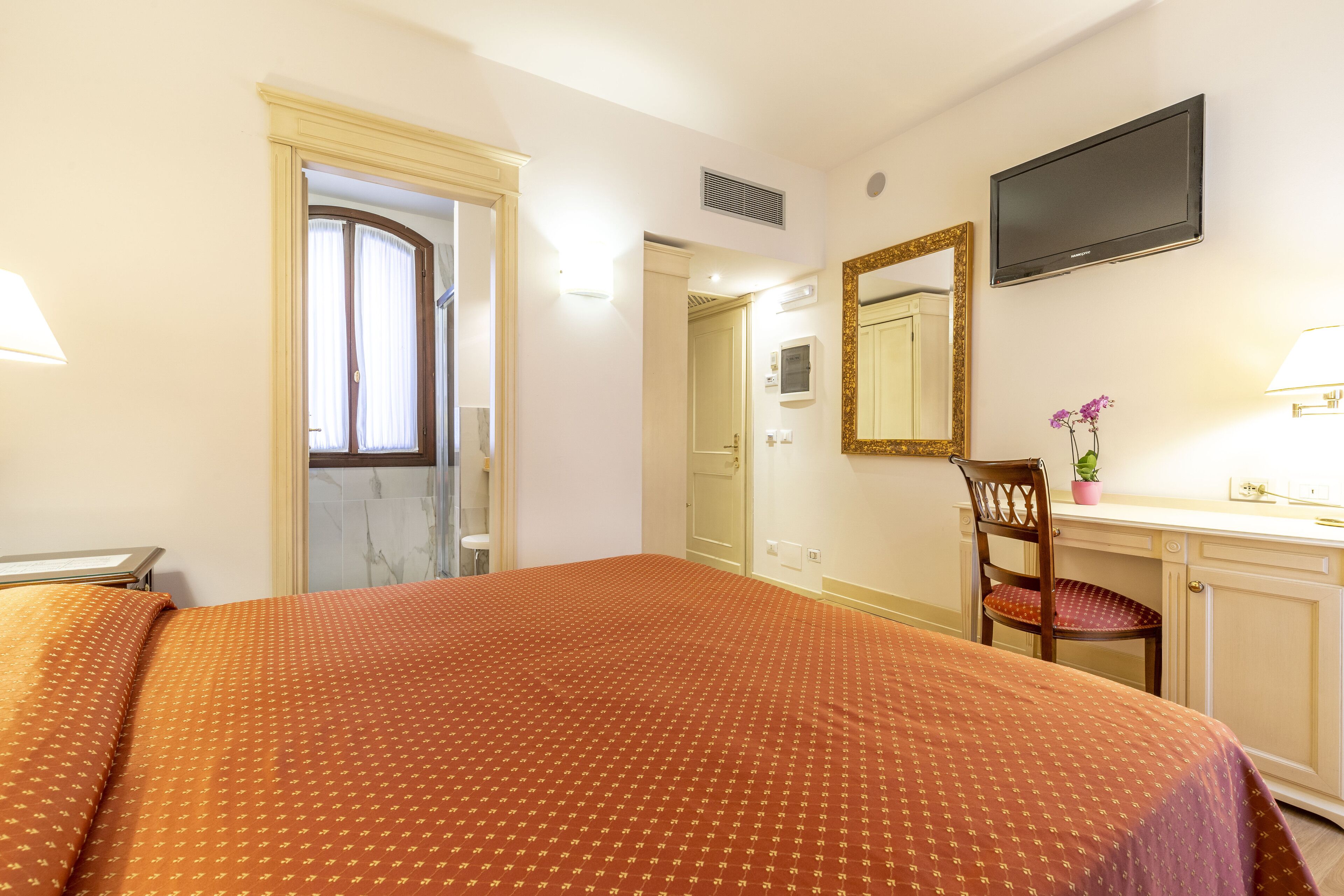 Habitación de hotel con cama, escritorio con silla, televisor y baño visible a través de una puerta abierta.