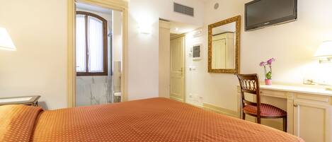 Habitación de hotel con cama, escritorio con silla, televisor y baño visible a través de una puerta abierta.