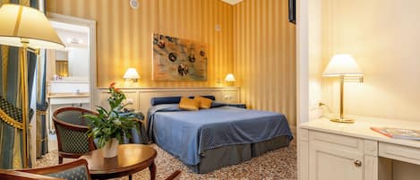 A hotel room with a bed, two bedside lamps, a desk, a chair, a small table with a plant, and a painting on the wall.