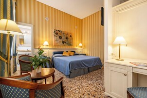 A hotel room with a bed, two bedside lamps, a desk, a chair, a small table with a plant, and a painting on the wall.