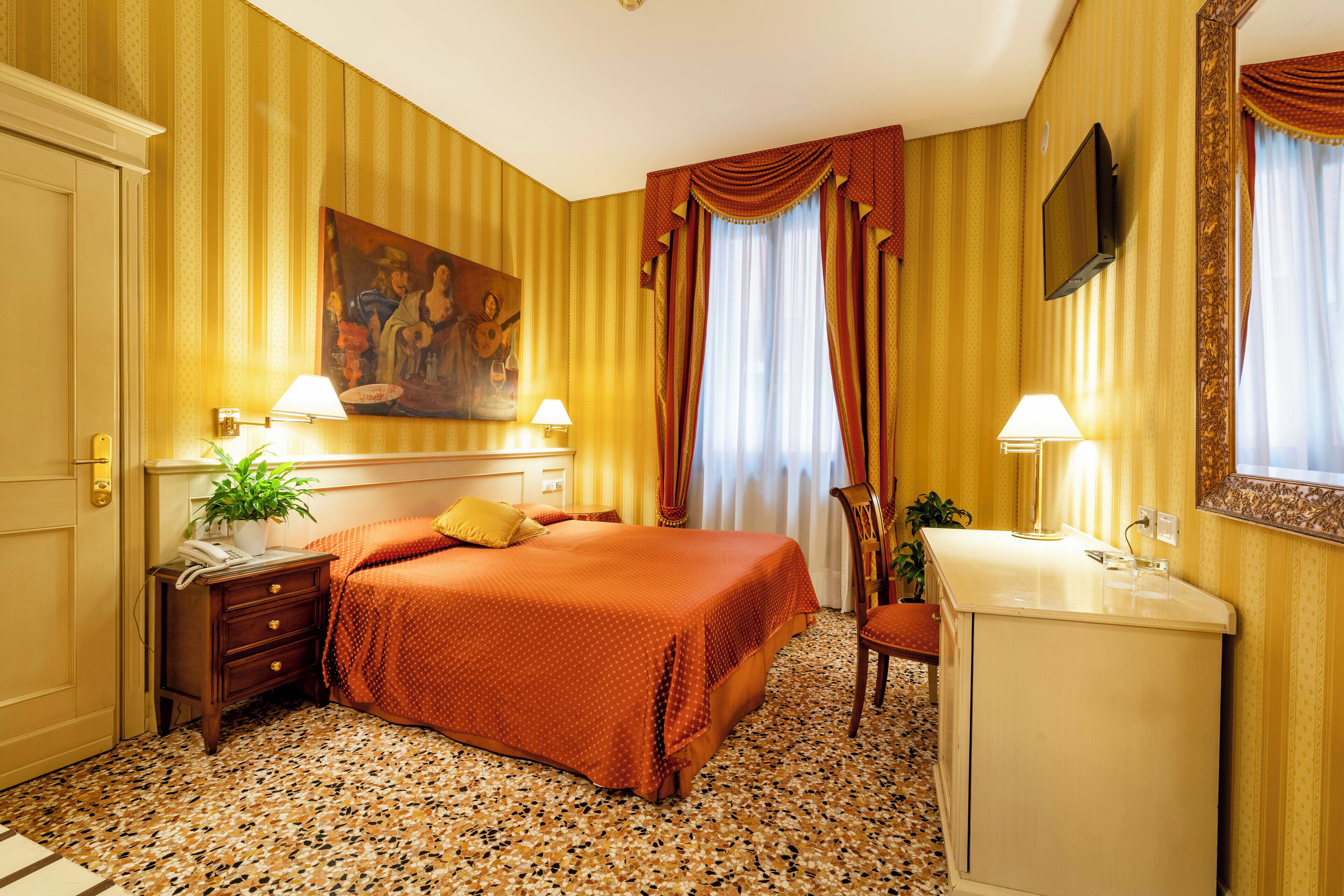 A hotel room with a bed, bedside table, lamp, desk, chair, and a painting on the wall.