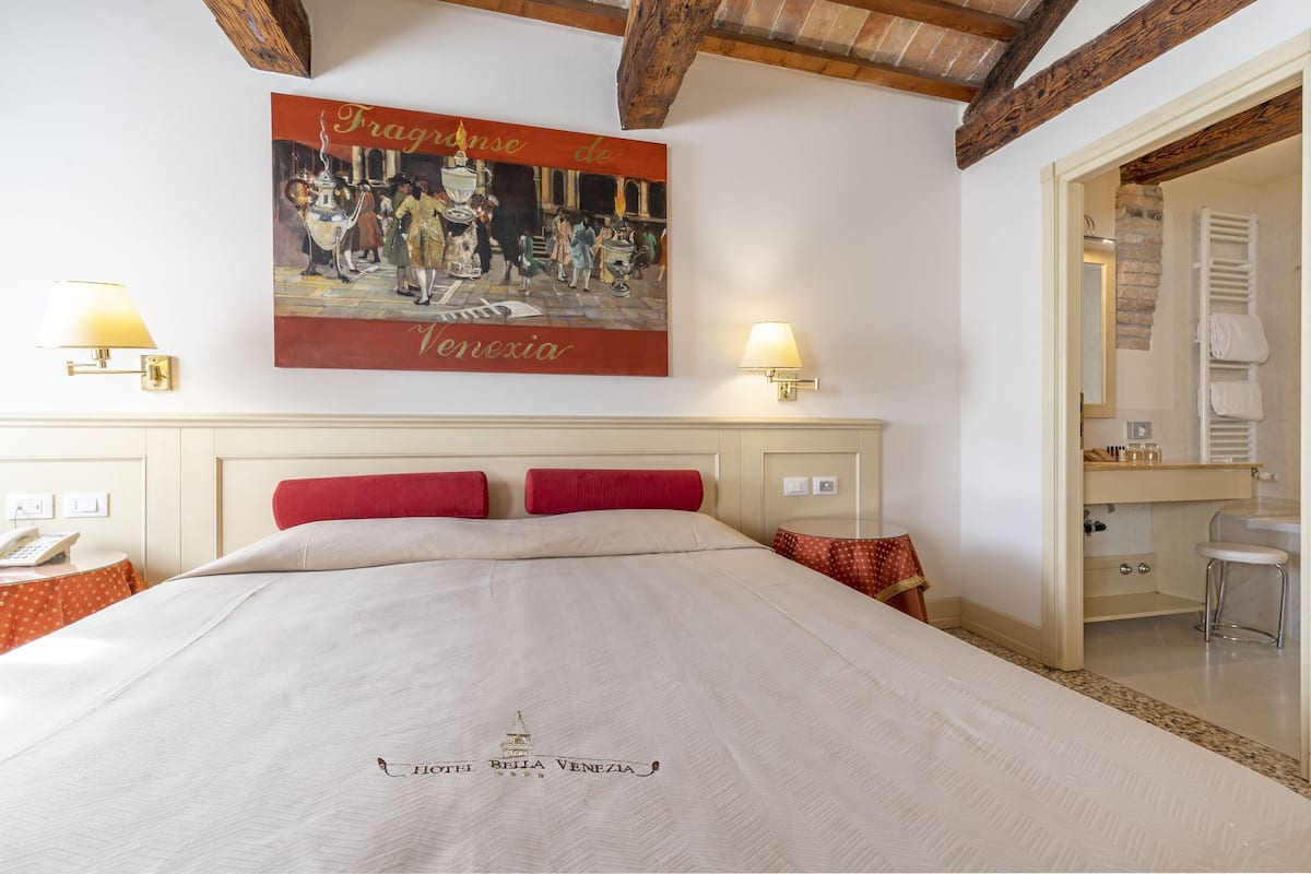 Ein Hotelzimmer mit einem Bett, roten Kissen, einem Gemälde, zwei Wandleuchten und einem durch eine offene Tür einsehbarer Baderaum.