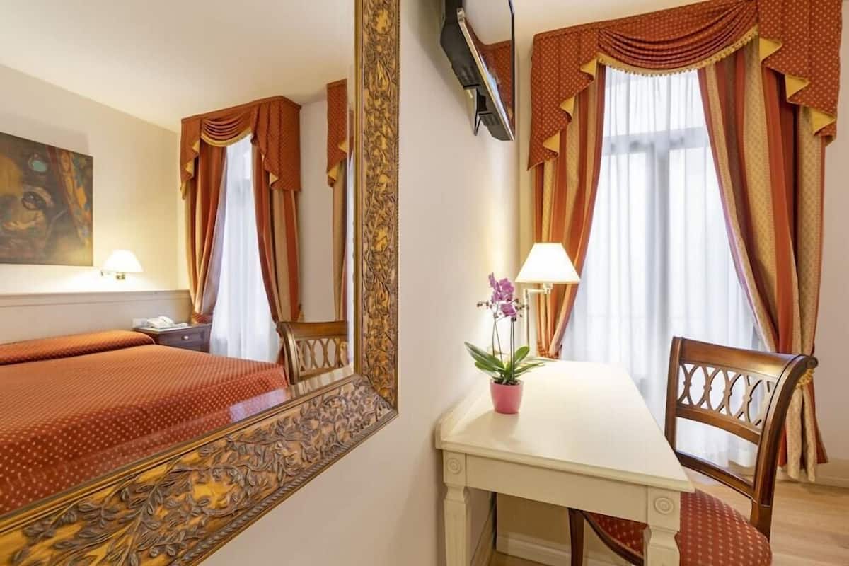 Ein Hotelzimmer mit einem Bett, einem Tisch mit Stuhl, einer Zimmerpflanze, einem Bild an der Wand und Vorhängen.