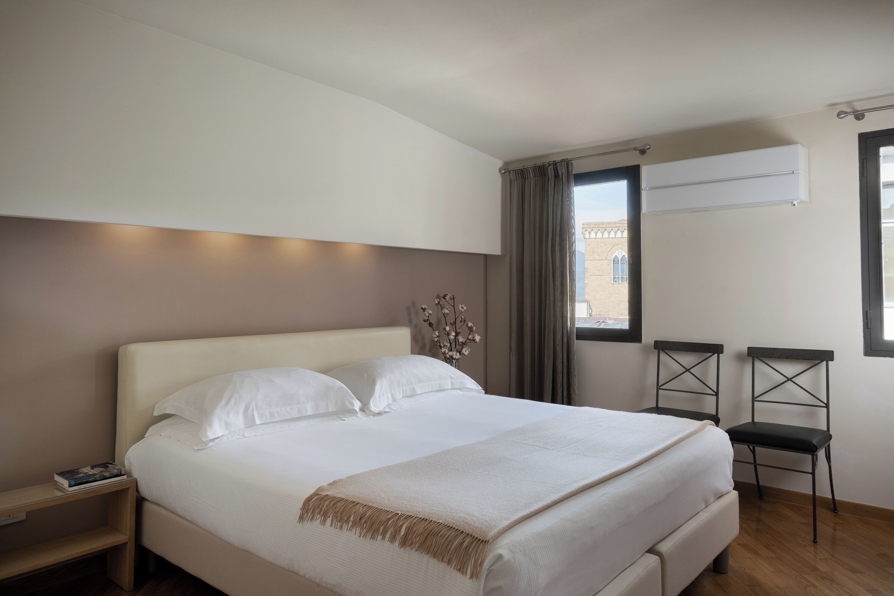 Una cama bien hecha con sábanas blancas, una mesita de noche con un libro y una ventana con cortinas.