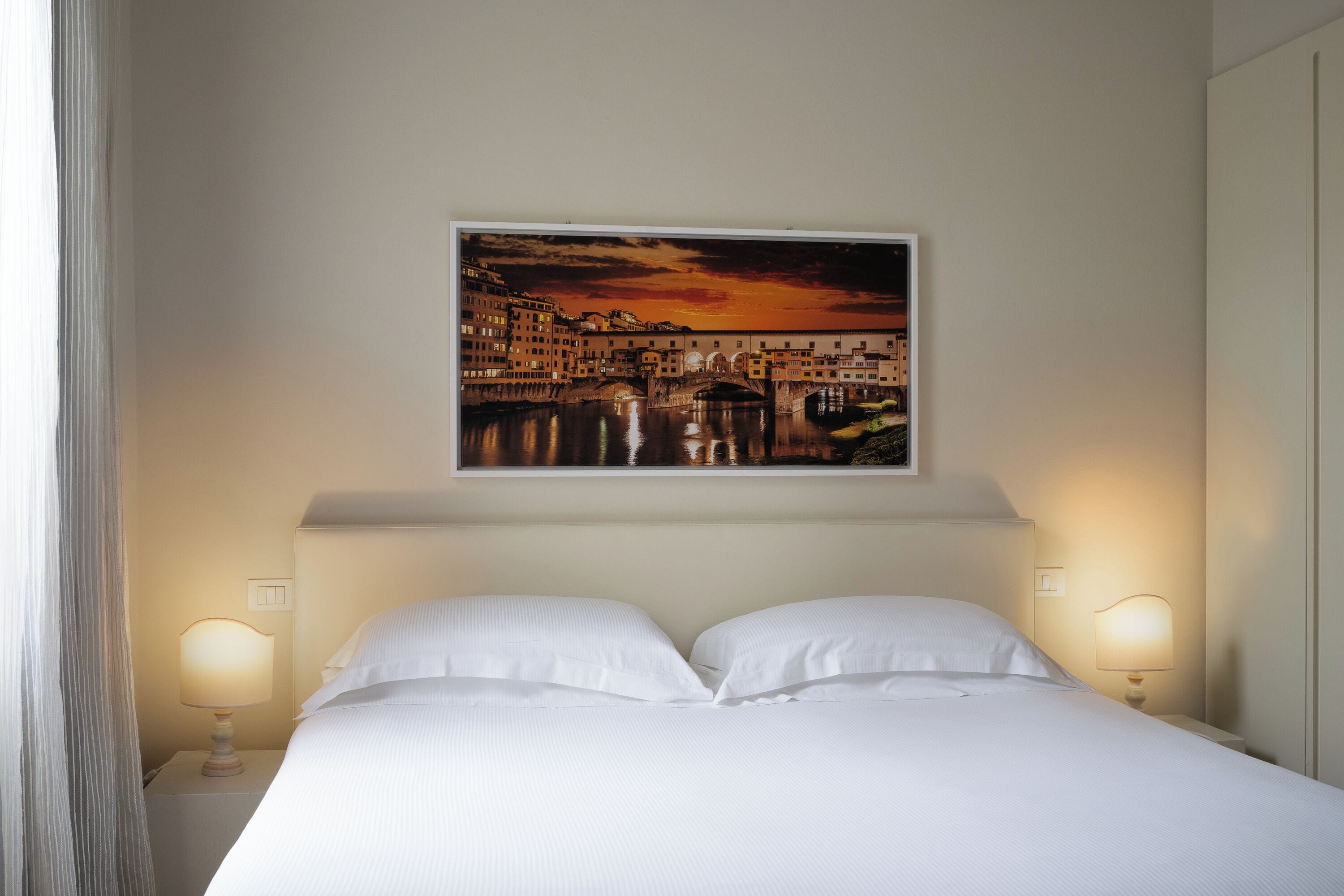 A hotel room with a bed, two bedside lamps, a painting of a cityscape, and a curtain on the left.