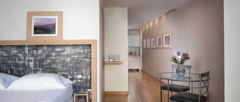 A bedroom with a bed, bedside table, and a small chair. There is a view of a kitchen and dining area with a table and chairs.
