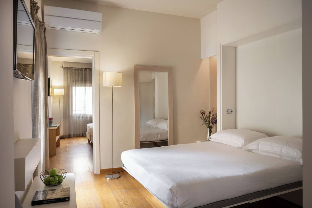 Une chambre d’hôtel avec un grand lit, une table de chevet avec une lampe, un miroir et une vue sur le couloir.