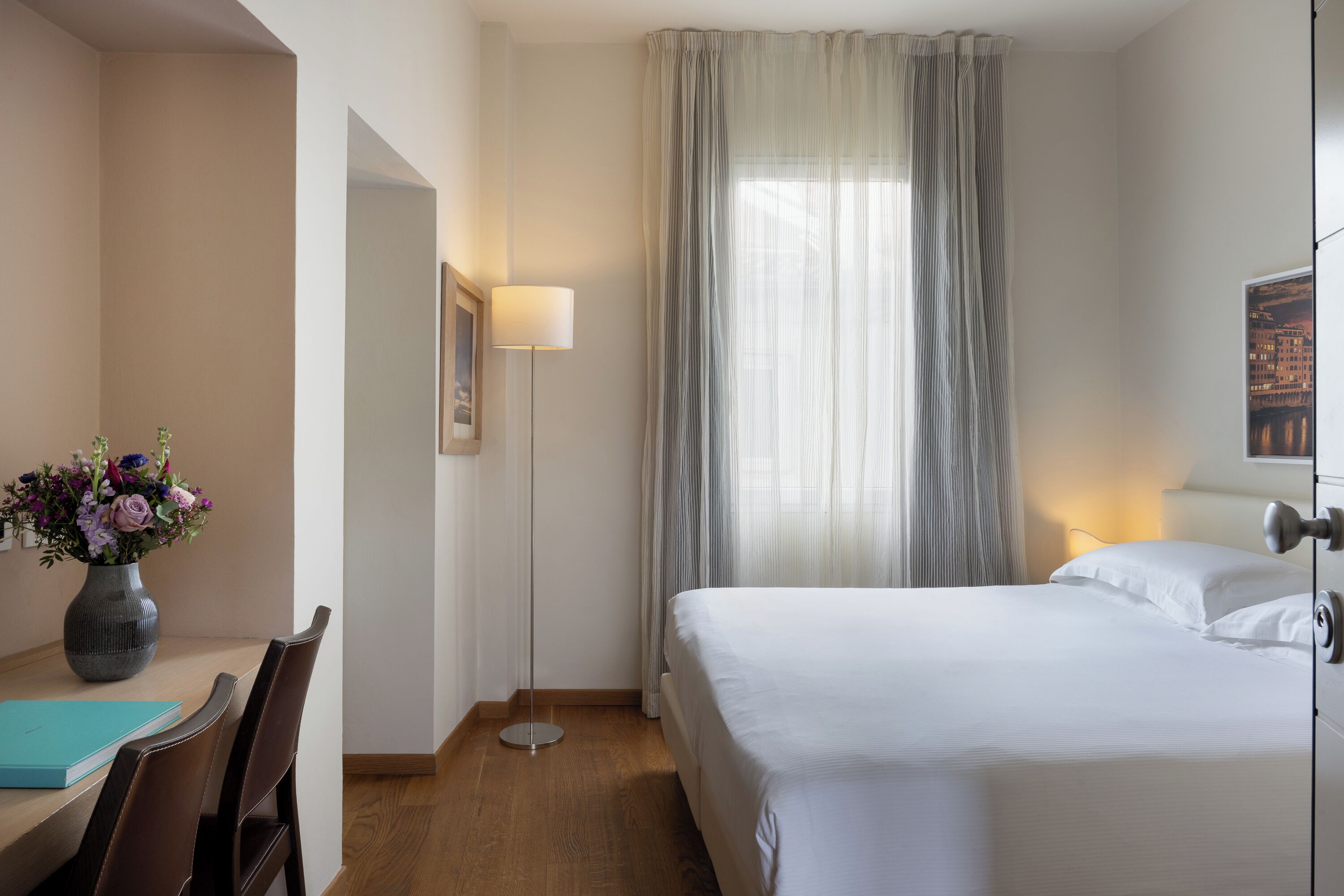 Et hotelværelse med en seng, et skrivebord med en blomstervase, en lampe og udsigt til en bygning gennem lette gardiner.