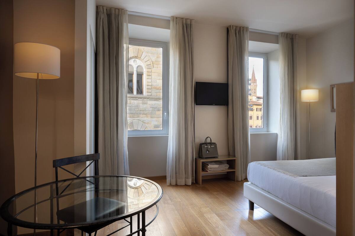 Une chambre d’hôtel avec un lit, un téléviseur fixé au mur, une table avec une chaise et une vue sur un immeuble par la fenêtre.