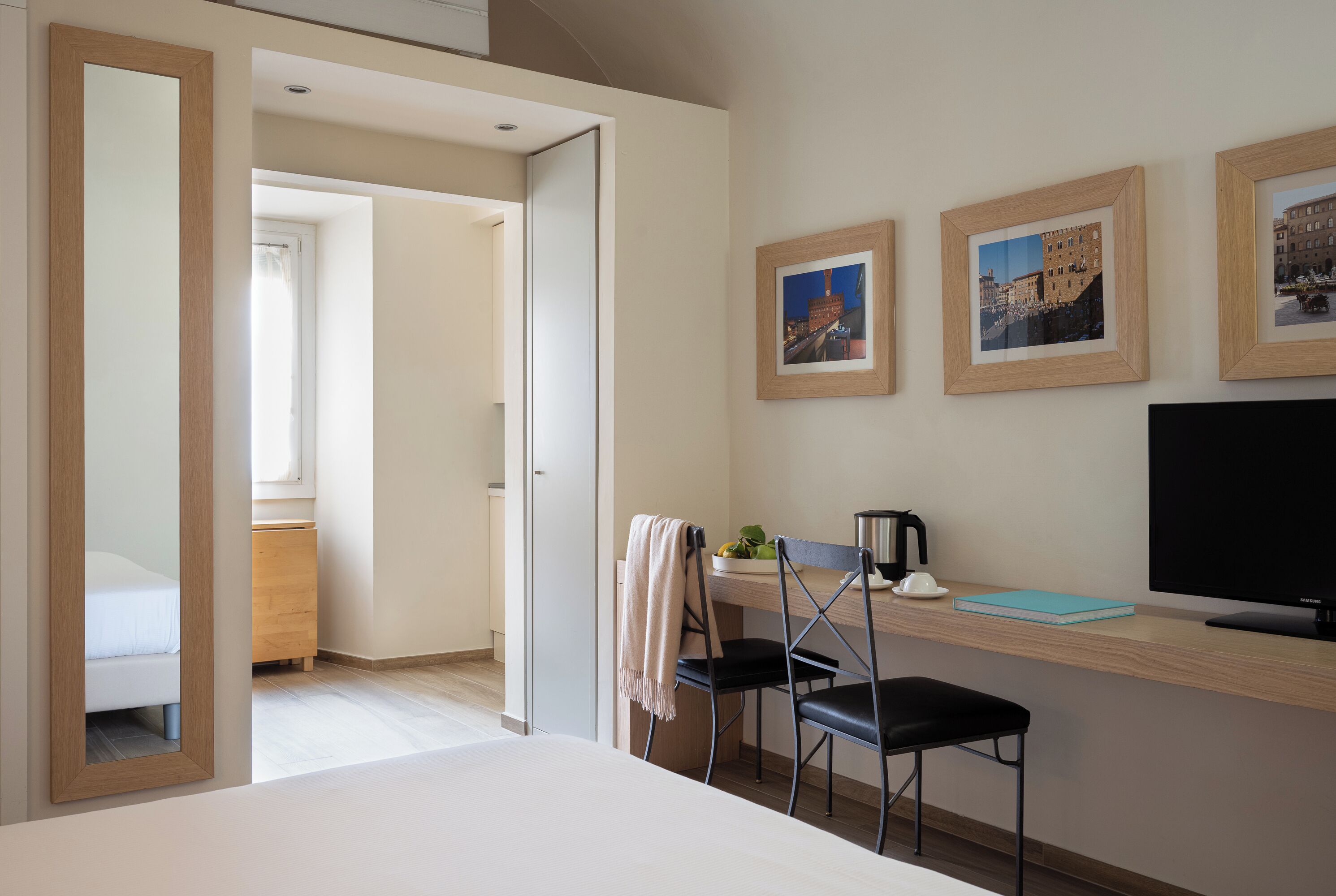 A hotel room with a bed, a desk with a chair, a TV, and framed pictures on the wall.