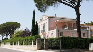 A pink two-story house with a balcony, surrounded by a stone wall and greenery.