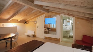 A cozy attic bedroom with a wooden ceiling, a dining table, a red chair, and a view into a bathroom through a glass door.