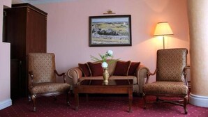 A room with a wooden coffee table, two patterned armchairs, a sofa with red cushions, a framed landscape painting on the wall, and a standing lamp. - Stone Bridge Hotel (Skopje)