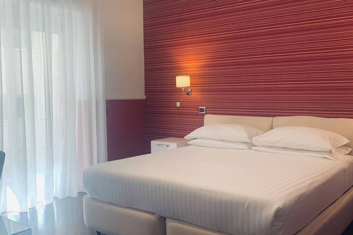 A hotel room with a large bed, a wooden headboard, a bedside table, and a window with sheer curtains.