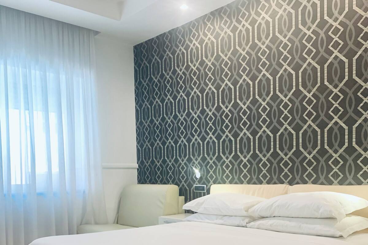 A neatly made bed with white linens in a room with wooden flooring, a patterned wall, and a window with sheer curtains.