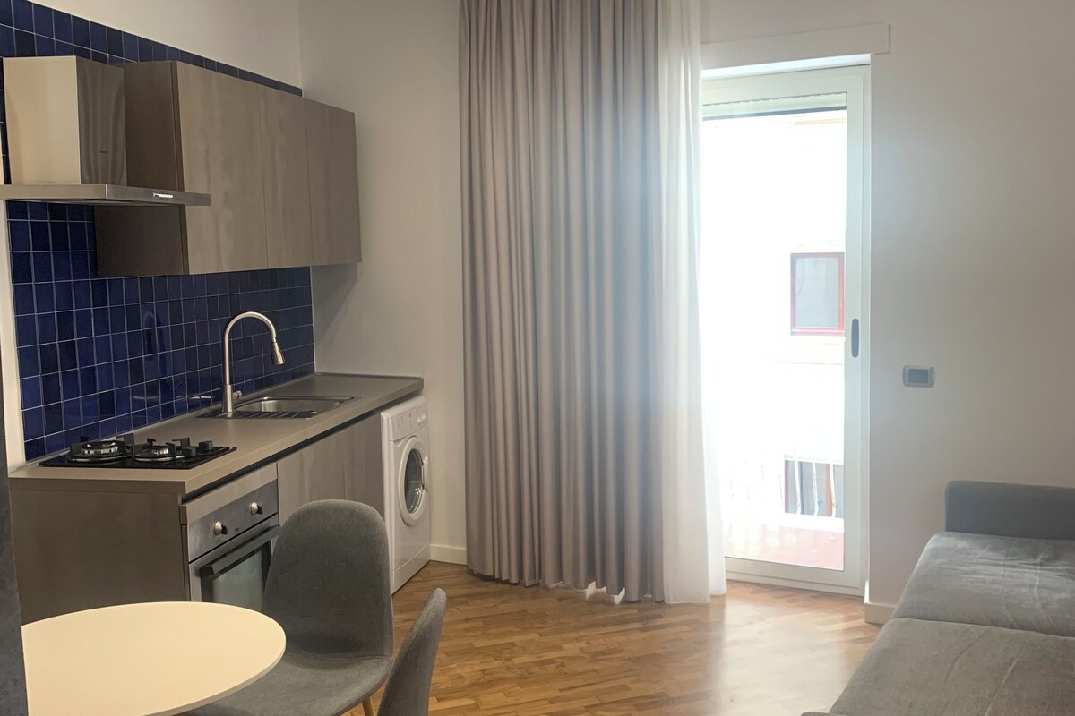 A compact living space with a kitchenette, a dining area with a table and chairs, a sofa, and a sliding glass door leading to an outdoor area.