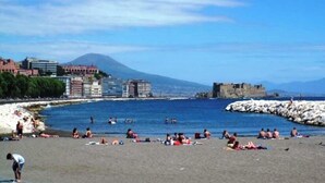 A beach with people sunbathing and swimming, buildings in the background, and a rocky outcrop.