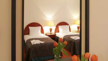 A hotel room with a large mirror reflecting two beds, a nightstand with lamps, and a vase with flowers.