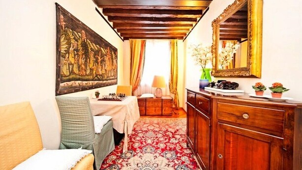 A cozy room with a wooden ceiling, a large painting, a table with a lamp, a mirror, and a dresser with small plants. - Residenza Giardini (Venice)