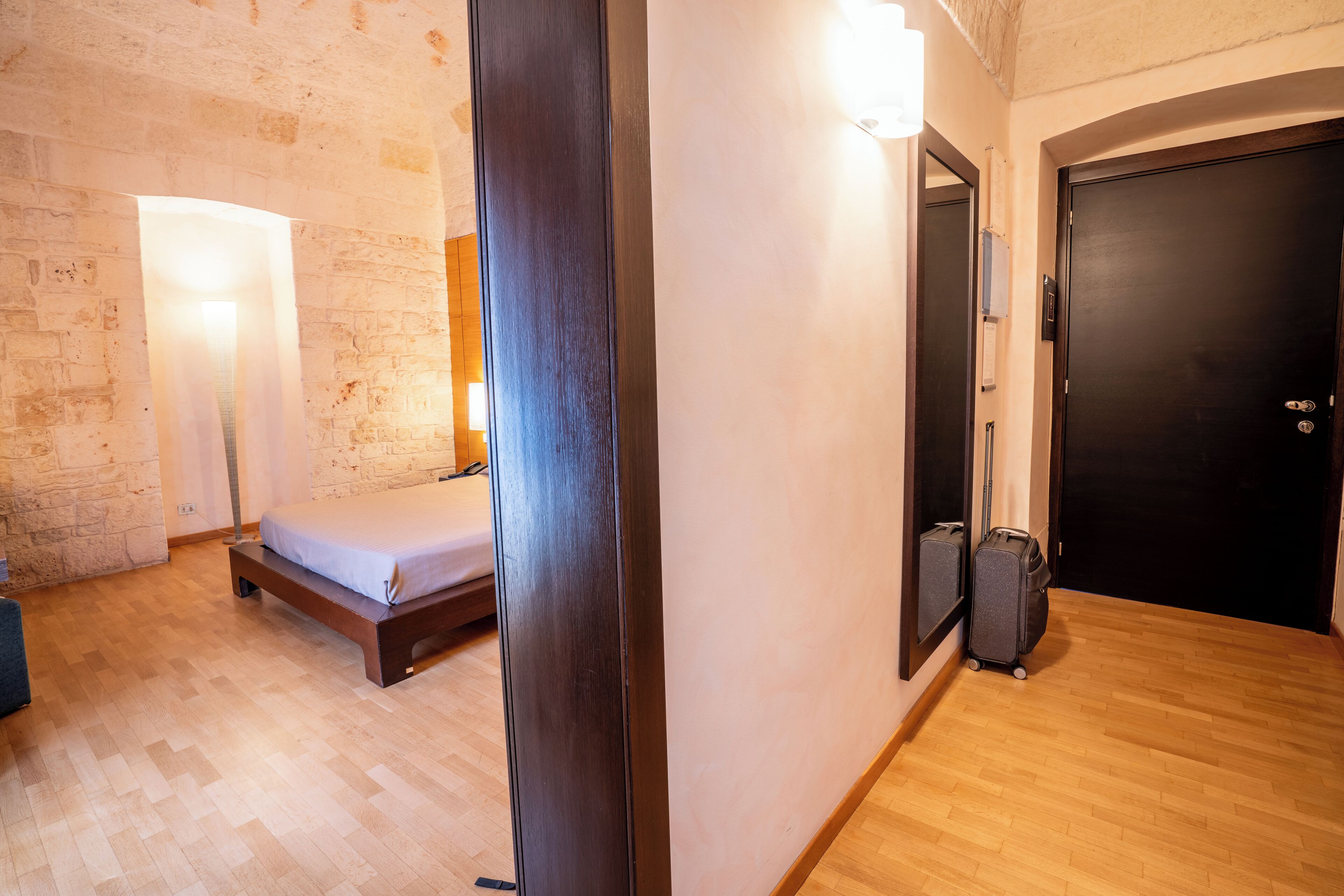 A room with a bed, a bench, a wooden floor, a wall with a light fixture, and a door.