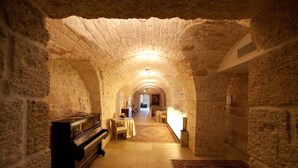 Un pasillo de paredes de piedra con un piano, mesas y sillas, que conduce a una habitación con cama.