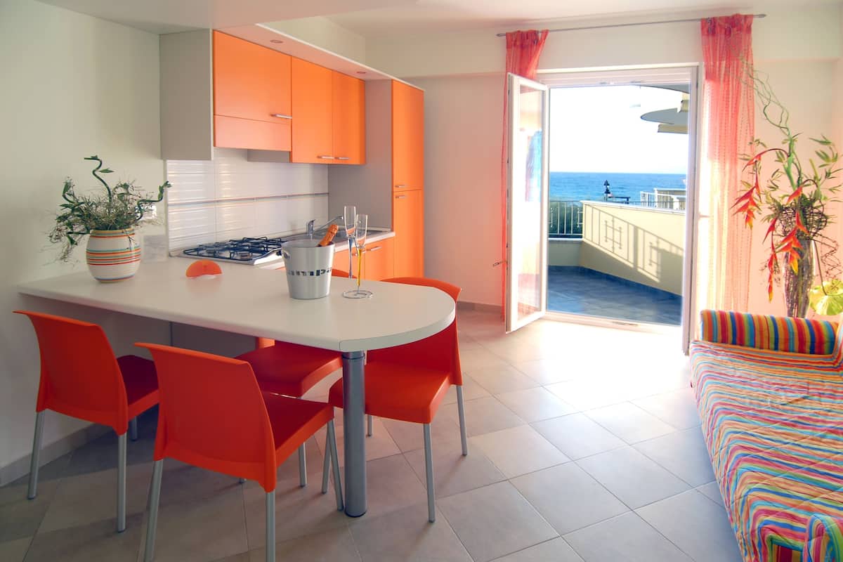Eine moderne Küche mit orangenen Schränken, einem Esstisch mit vier Stühlen und Meerblick durch eine offene Tür.