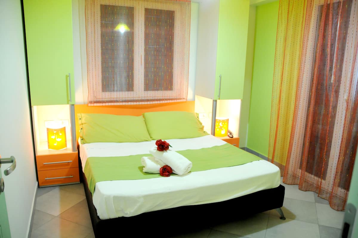 Ein ordentlich gemachtes Bett mit grüner Bettwäsche und zwei roten Rosen, ein Fenster mit durchscheinenden Vorhängen und zwei Nachttischlampen.