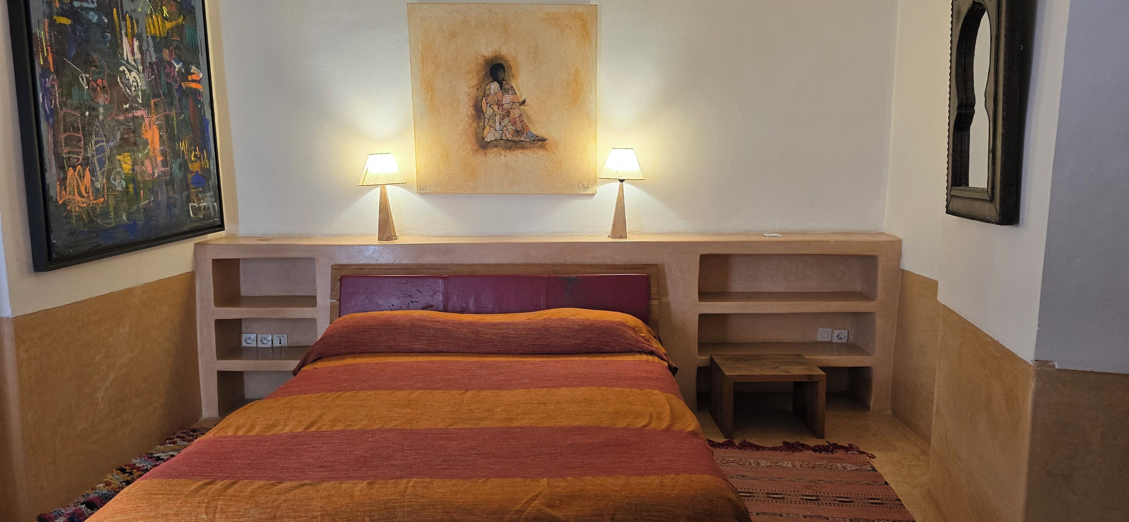 A bedroom with a bed, two bedside lamps, a painting above the bed, and a small table beside the bed.