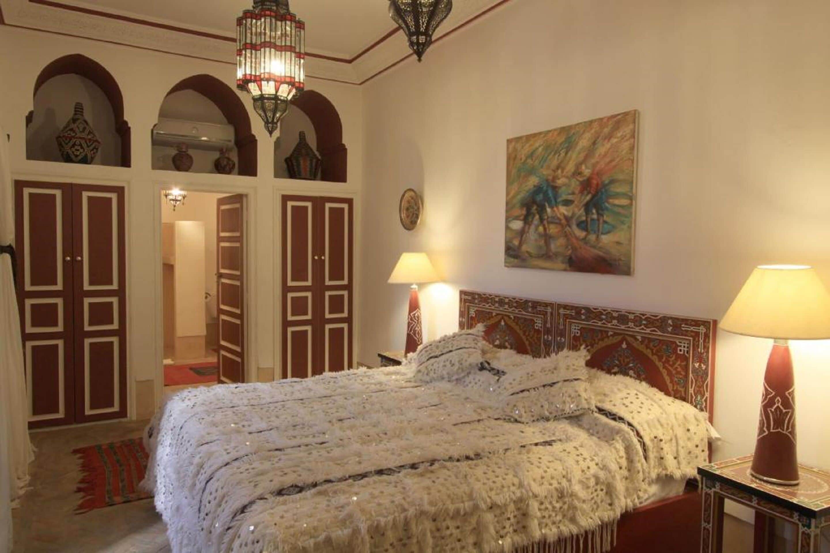A bedroom with a large bed, two bedside lamps, a chandelier, and a painting on the wall.