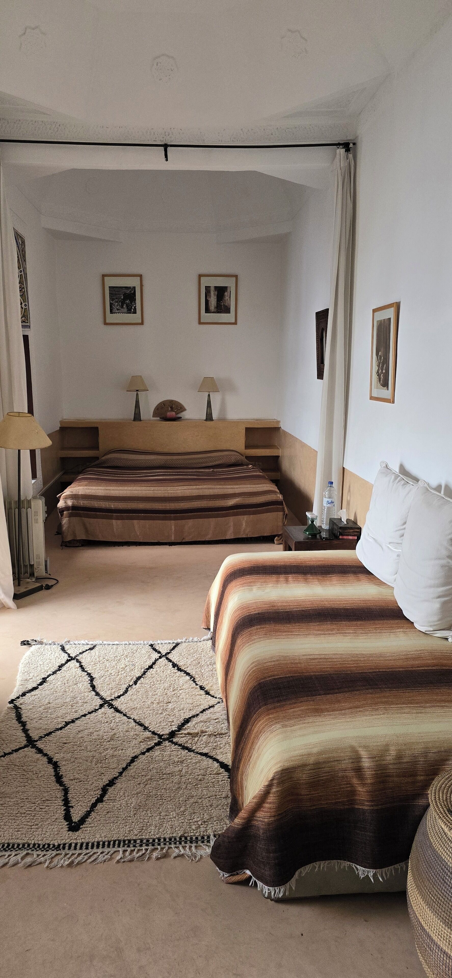 Habitación de hotel con una cama grande, dos camas más pequeñas, una mesita de noche, una lámpara y una mesita auxiliar con una botella.