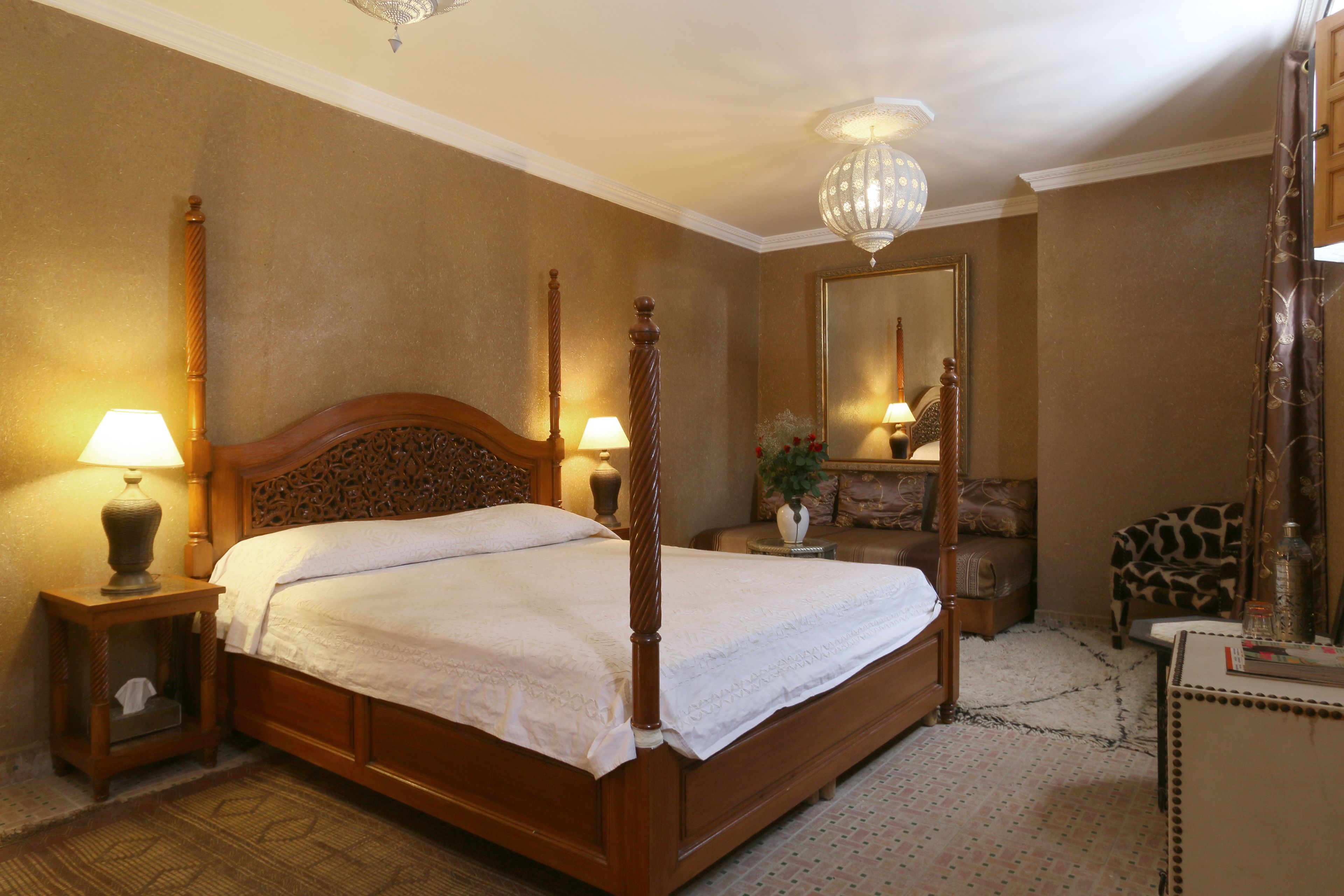 A four-poster bed with a wooden headboard, two bedside tables with lamps, a mirror, a giraffe-patterned chair, and a small table with a vase and bottles.