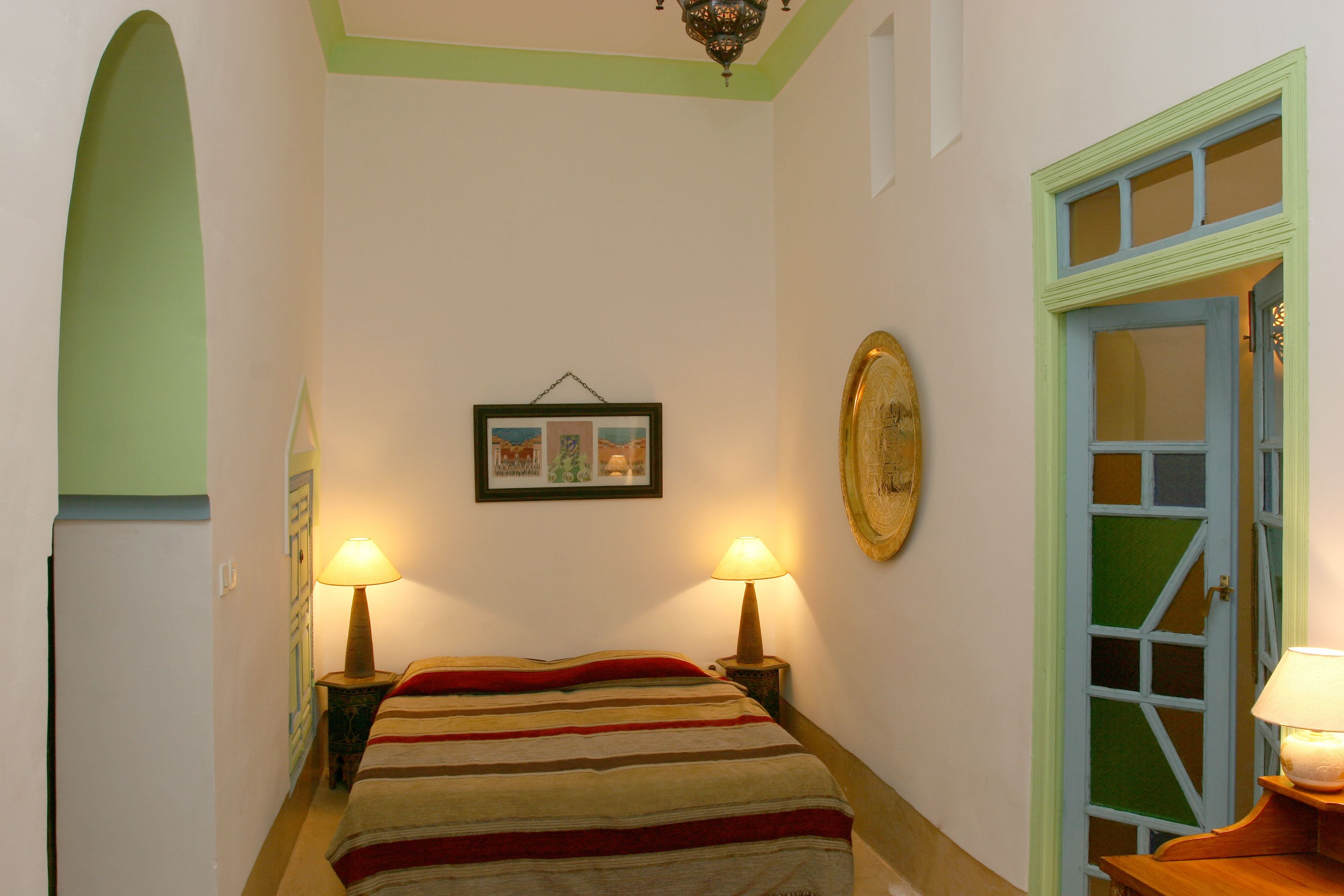 Une petite pièce avec un lit, deux lampes de chevet, un tableau encadré au mur et une porte à cadre vert.