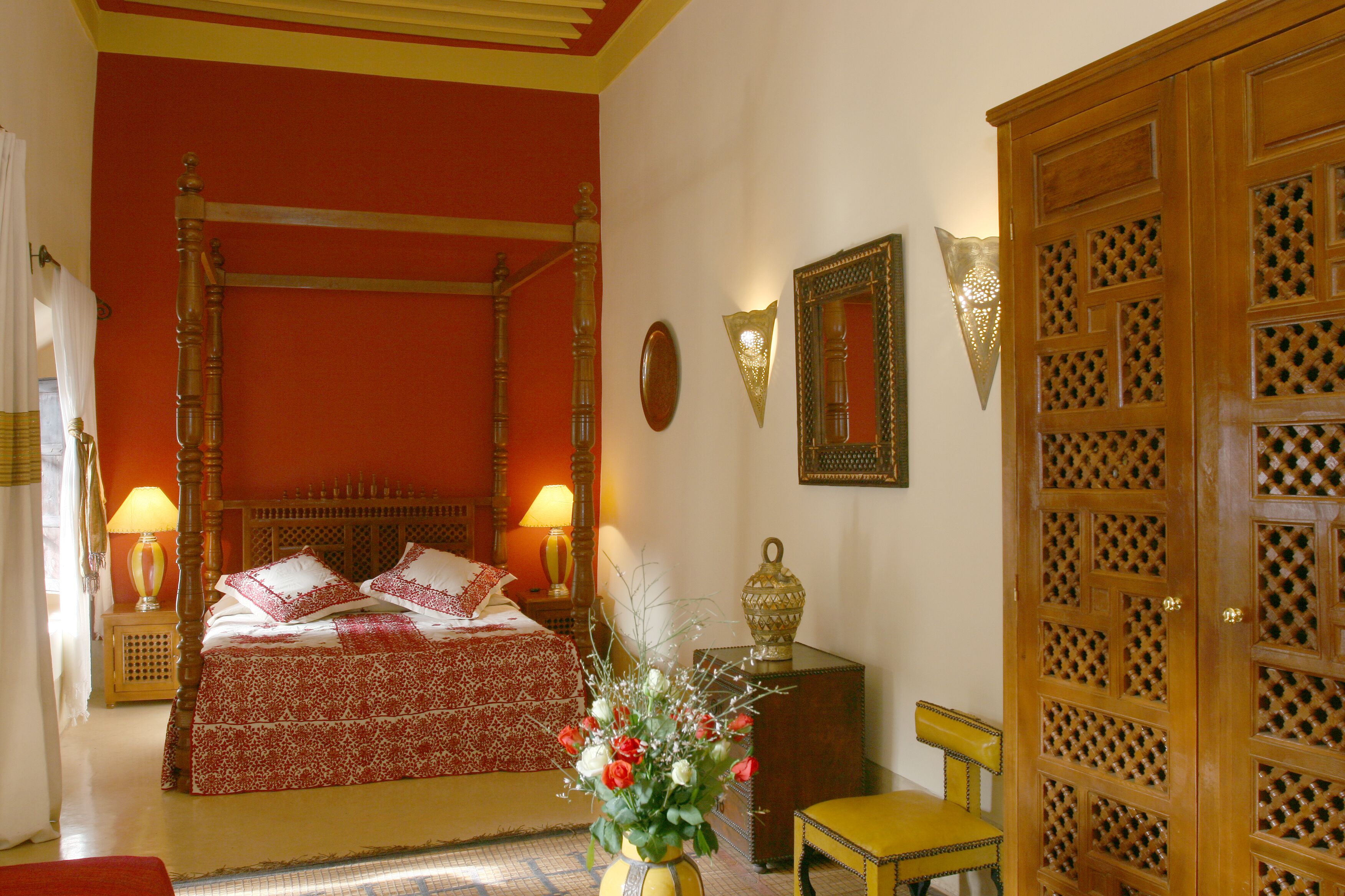 A four-poster bed with a red and white bedspread, a wooden nightstand with a lamp, a yellow chair, and a wooden wardrobe with a lattice design.