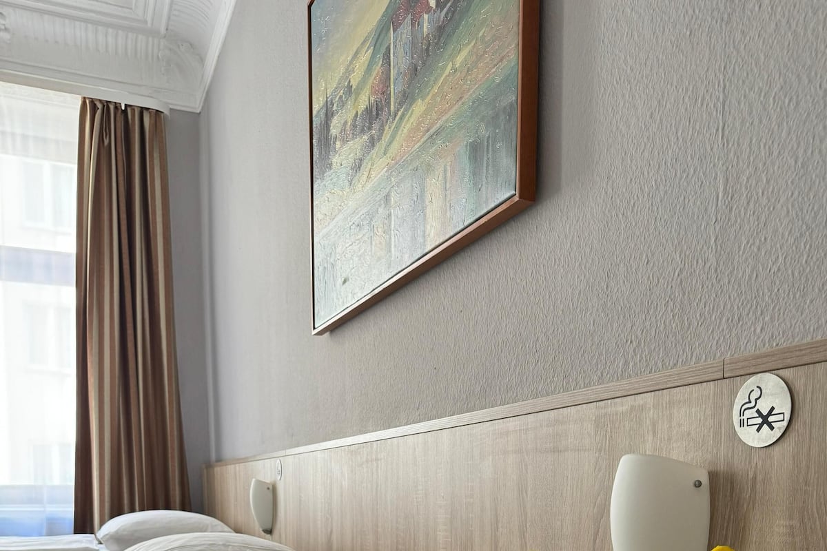 Une chambre d’hôtel avec un grand lit, du linge de lit blanc, un tableau au mur, une tête de lit en bois, un vase blanc avec des fleurs jaunes et un panneau indiquant qu’il est interdit de fumer.