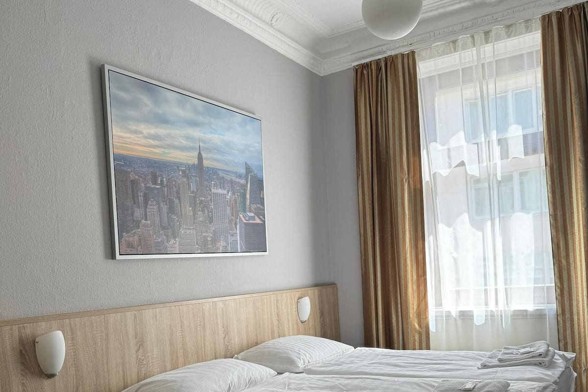 Une chambre d’hôtel avec un grand lit, une table de chevet, une lampe fixée au mur, un tableau représentant un paysage urbain et une fenêtre avec des rideaux.