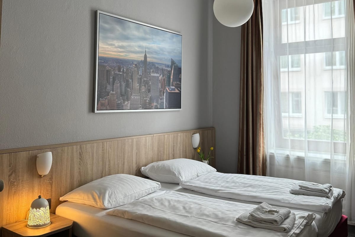 Une chambre d’hôtel avec deux lits, un tableau représentant un paysage urbain, un ventilateur de plafond et une fenêtre avec des rideaux.