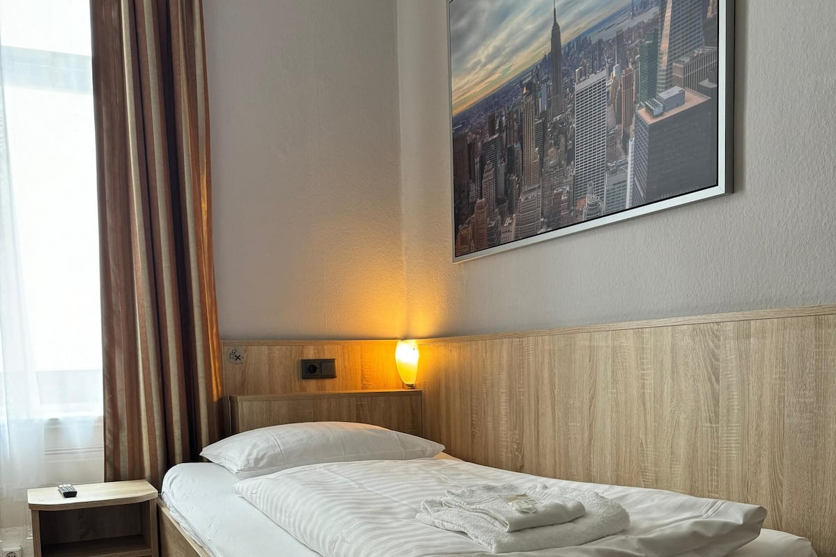 Une chambre d’hôtel avec un lit en bois, une table de chevet, une lampe et un tableau représentant un paysage urbain accroché au mur.