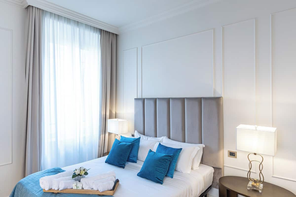 Una habitación de hotel con una cama grande, dos lámparas de noche, una mesita de noche y un ventanal con cortinas.