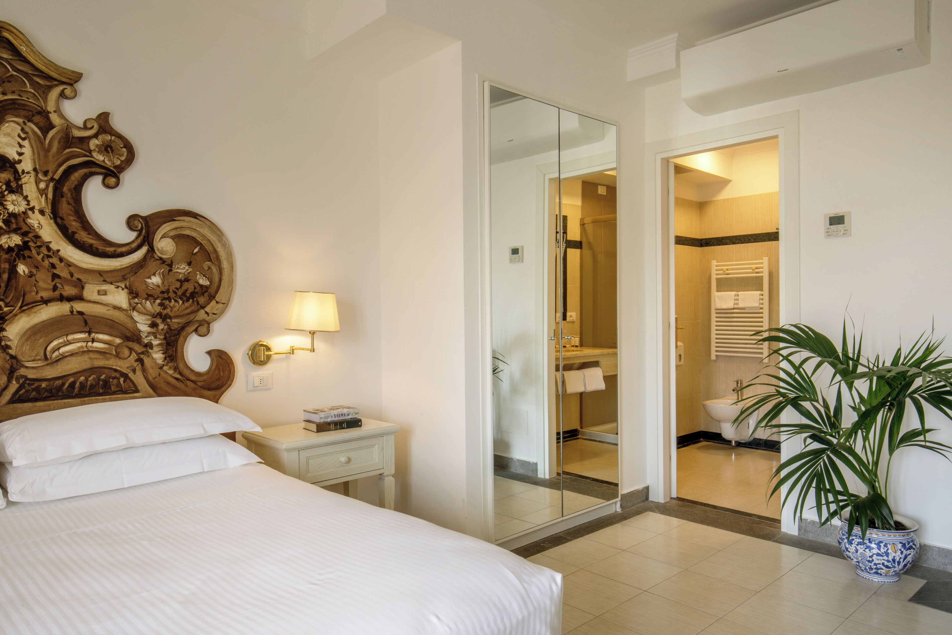 A hotel room with a large bed, a nightstand with a lamp, a mirror, a potted plant, and a bathroom visible through a glass door.