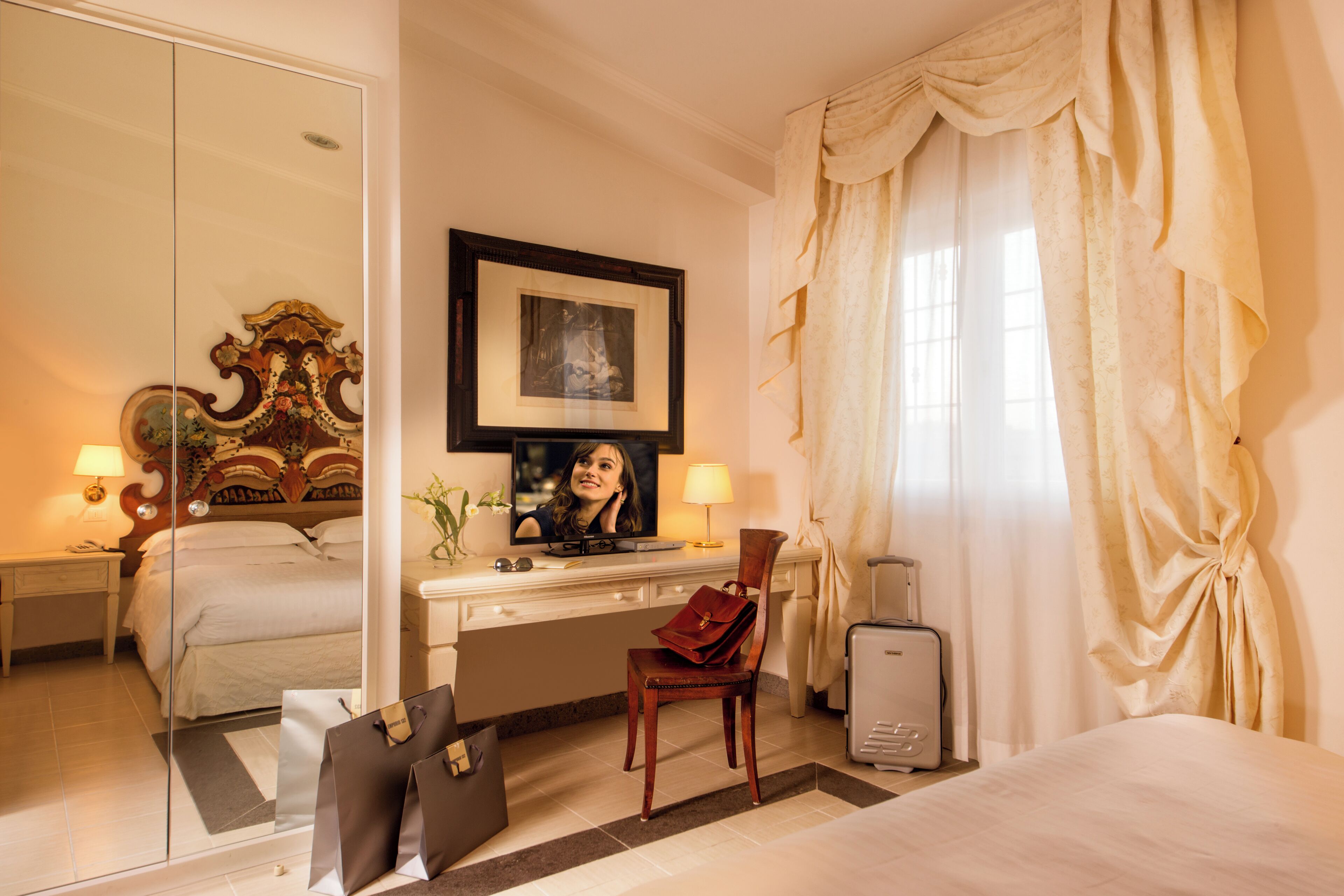 A hotel room with a bed, a desk, a chair, a suitcase, a television, and a mirror.