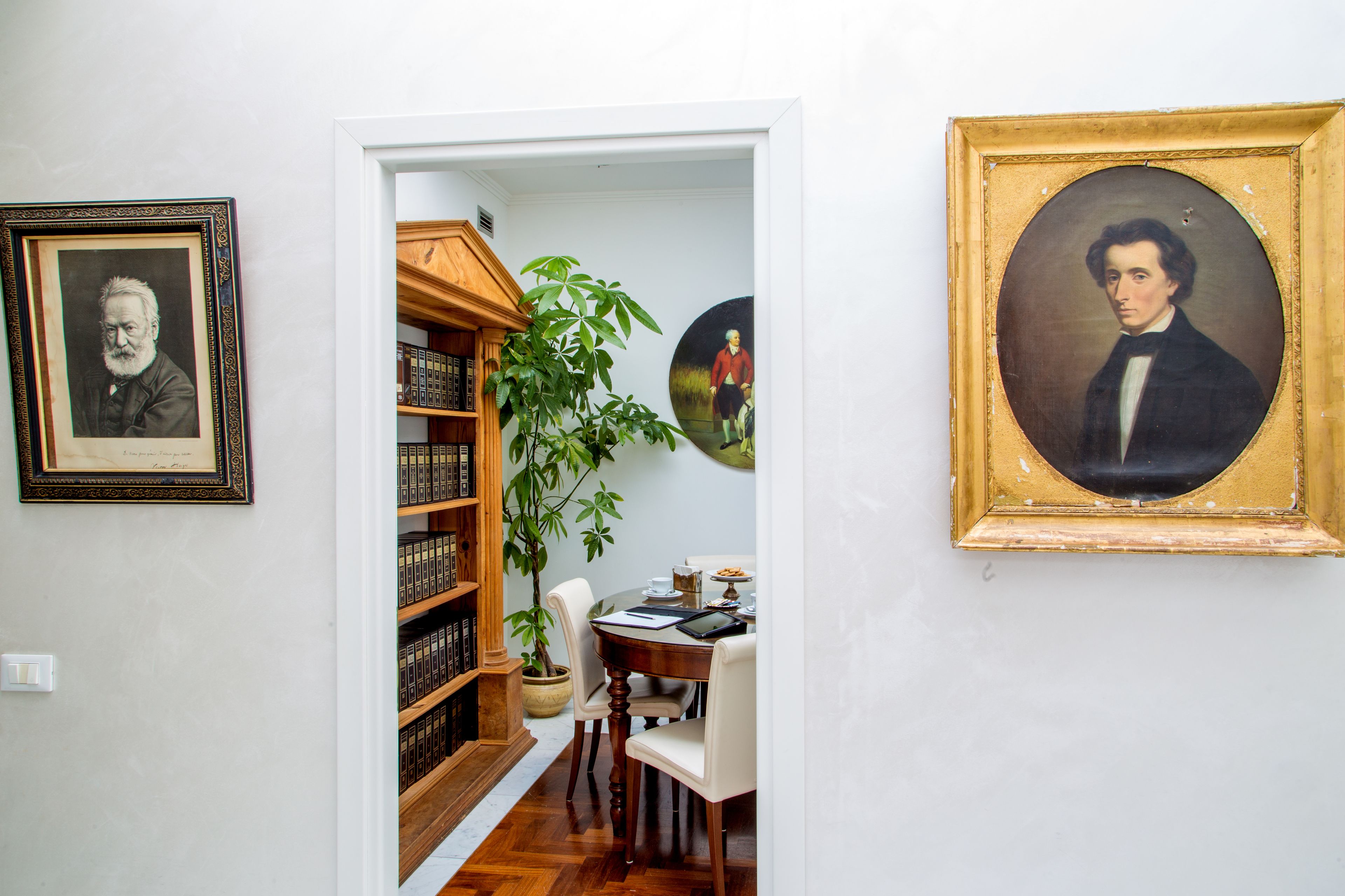 A room with a wooden bookshelf, a dining table with chairs, and framed portraits on the walls.