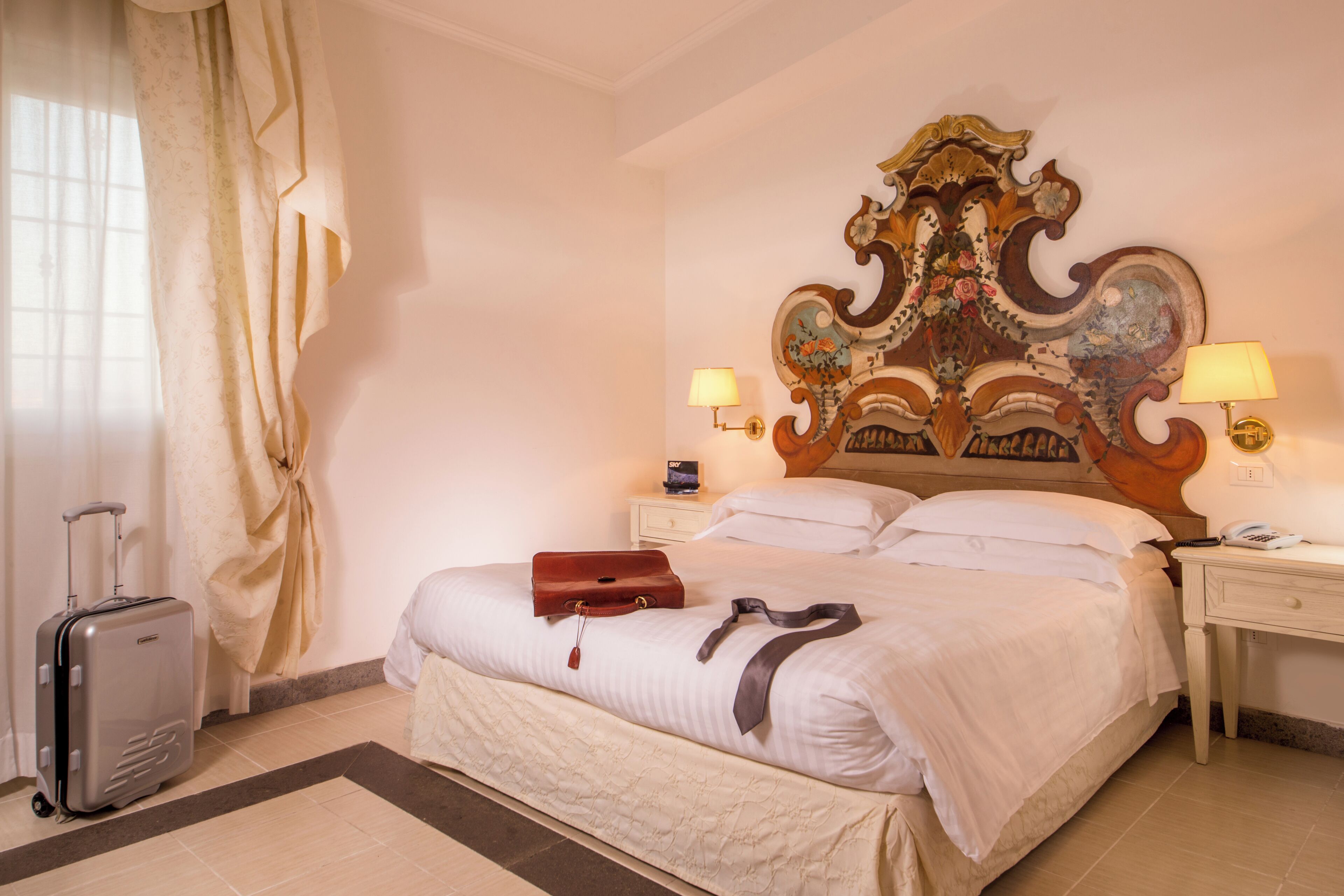 A hotel room with a large bed, a nightstand with a lamp, a suitcase, and a decorative headboard.