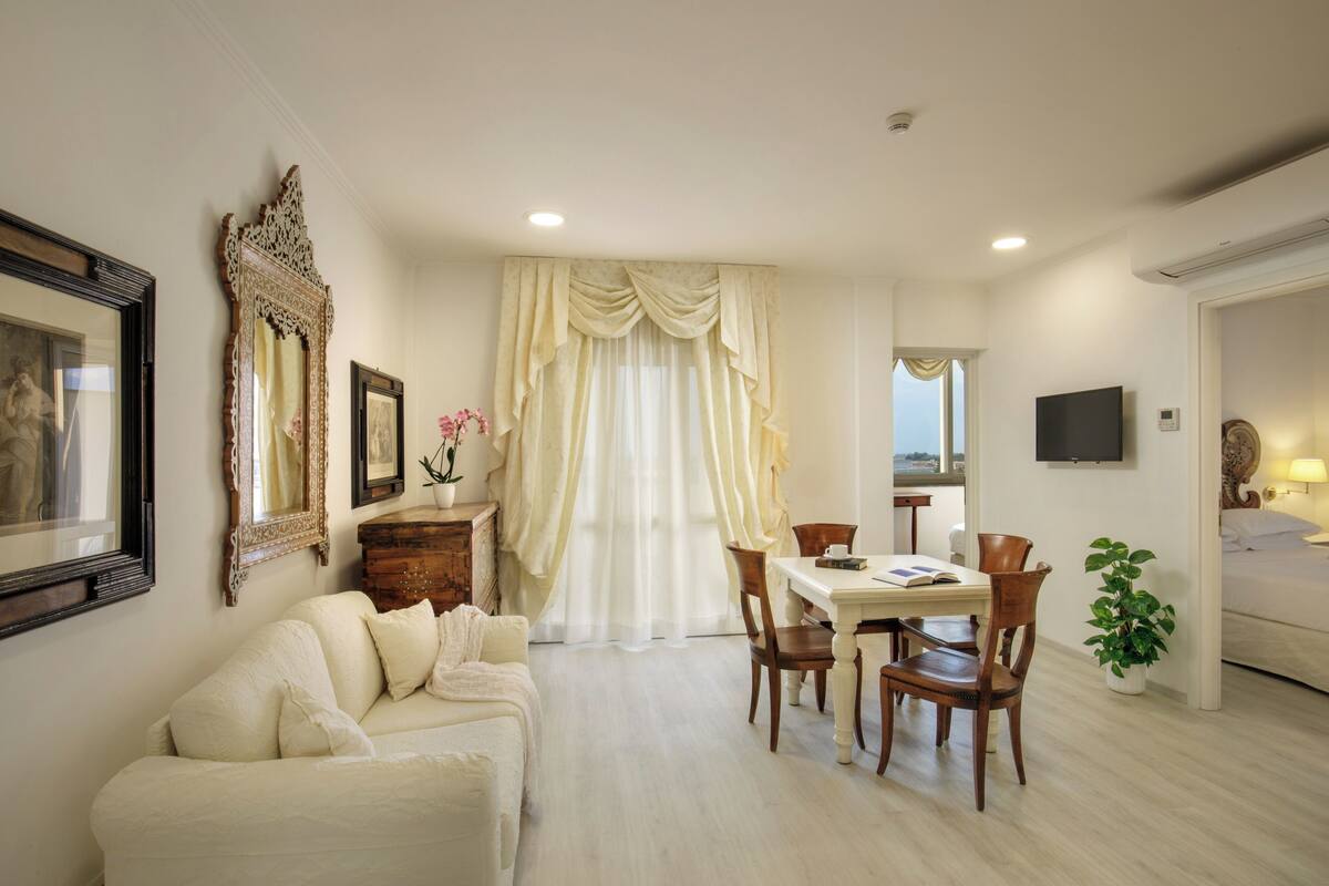 A hotel room with a dining area, a sofa, a TV, and a window with curtains.