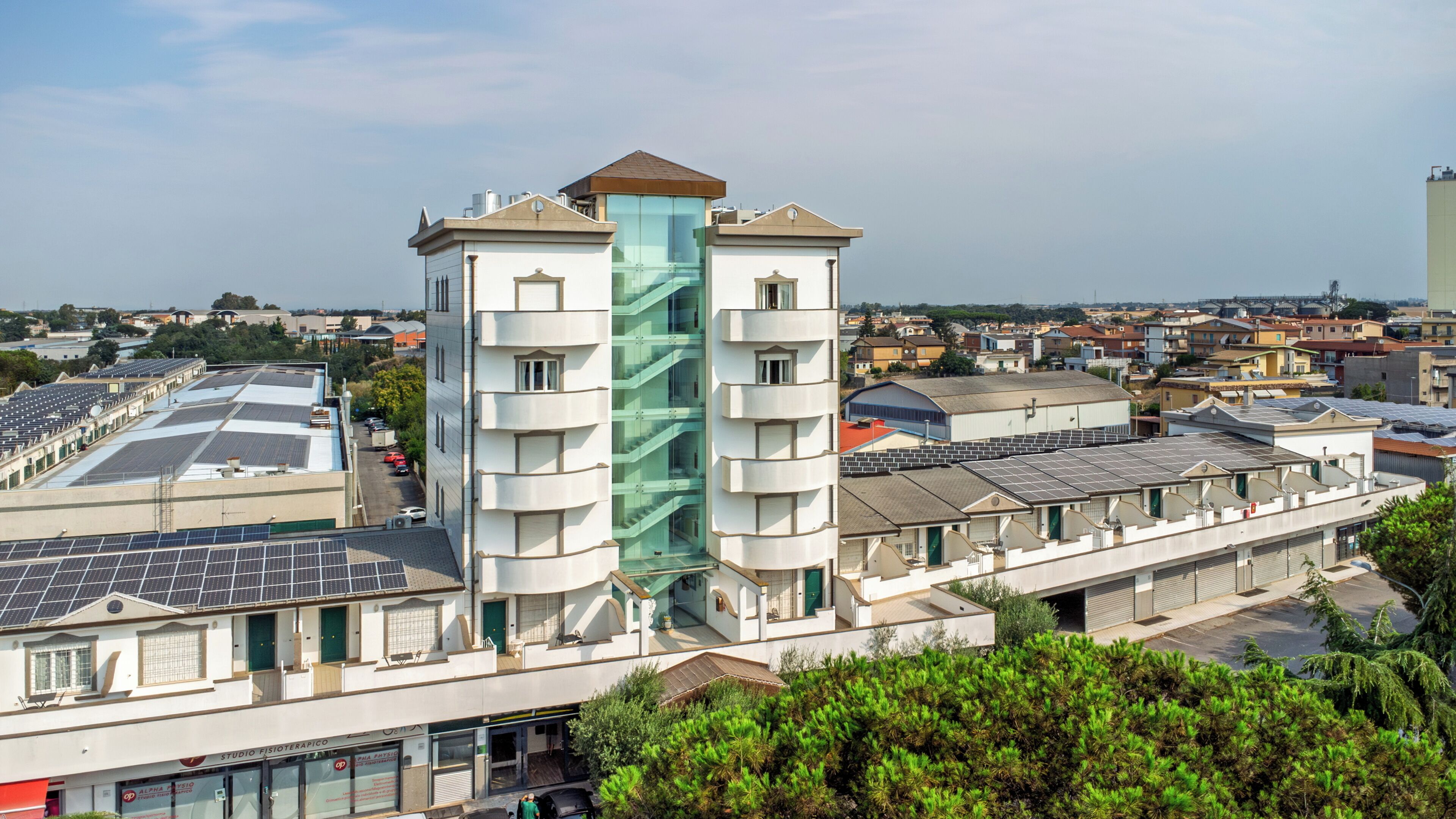 A multi-story building with a central glass elevator shaft, surrounded by residential buildings and solar panels on rooftops.