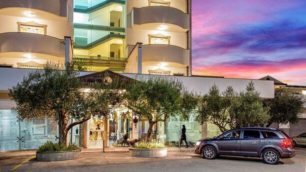 A multi-story hotel with a glass facade, outdoor seating area, and a parked car. - Arcom Palace (Pomezia)