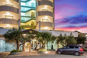 Un hôtel à plusieurs étages doté d’une façade en verre, d’un espace extérieur aménagé pour s’asseoir et d’une voiture garée.