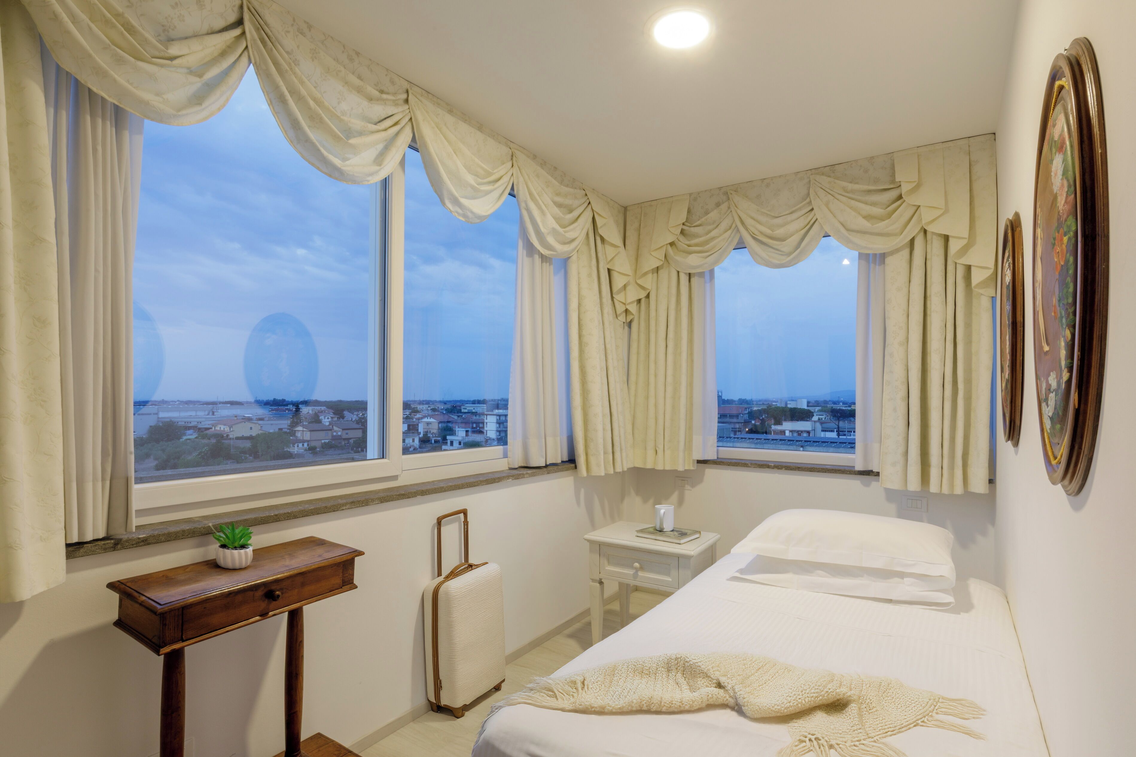 A hotel room with a bed, a bedside table with a lamp, a small table with a plant, a suitcase, and a view of the city through large windows.