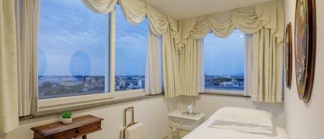 A hotel room with a bed, a bedside table with a lamp, a small table with a plant, a suitcase, and a view of the city through large windows.