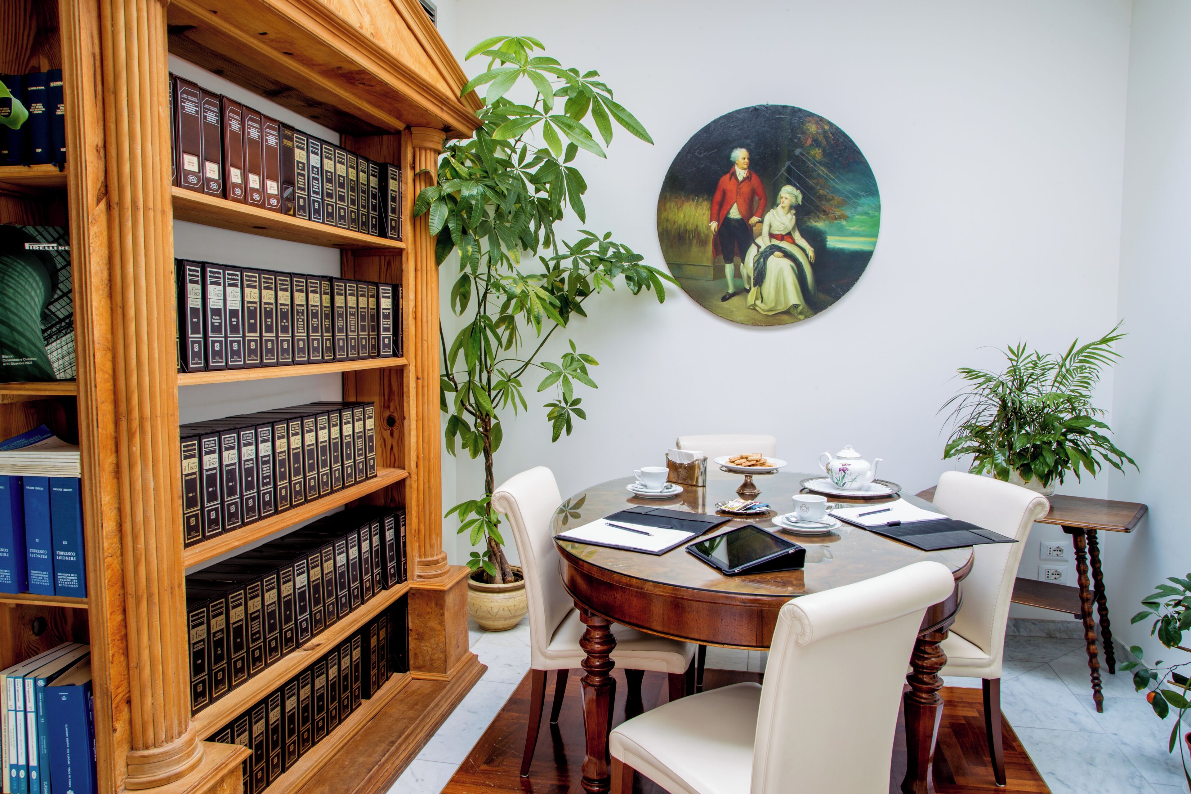 A dining area with a round table, chairs, a bookshelf, and a painting on the wall.