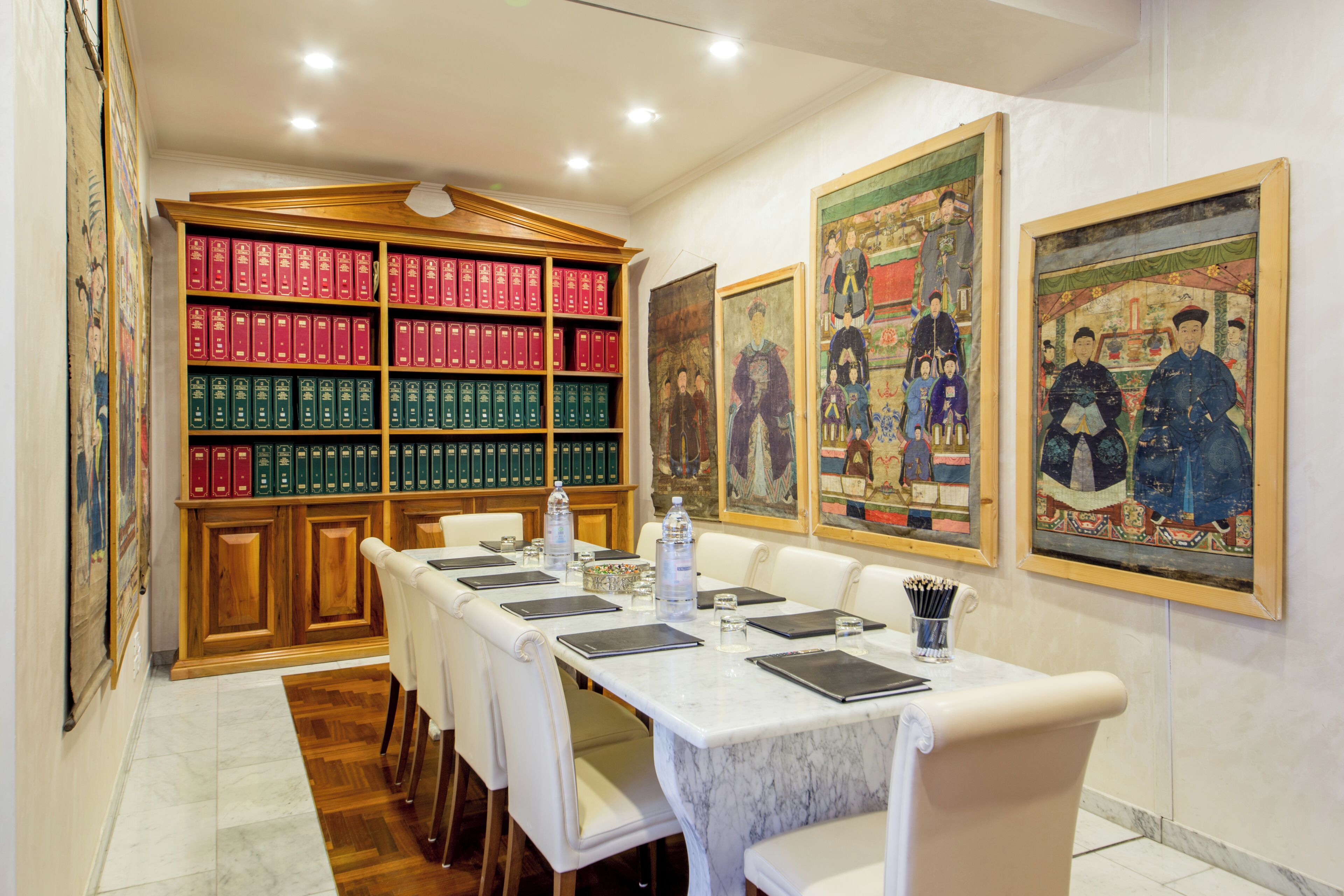 A conference room with a long table, white chairs, a bookshelf with red and green books, and traditional paintings on the walls.